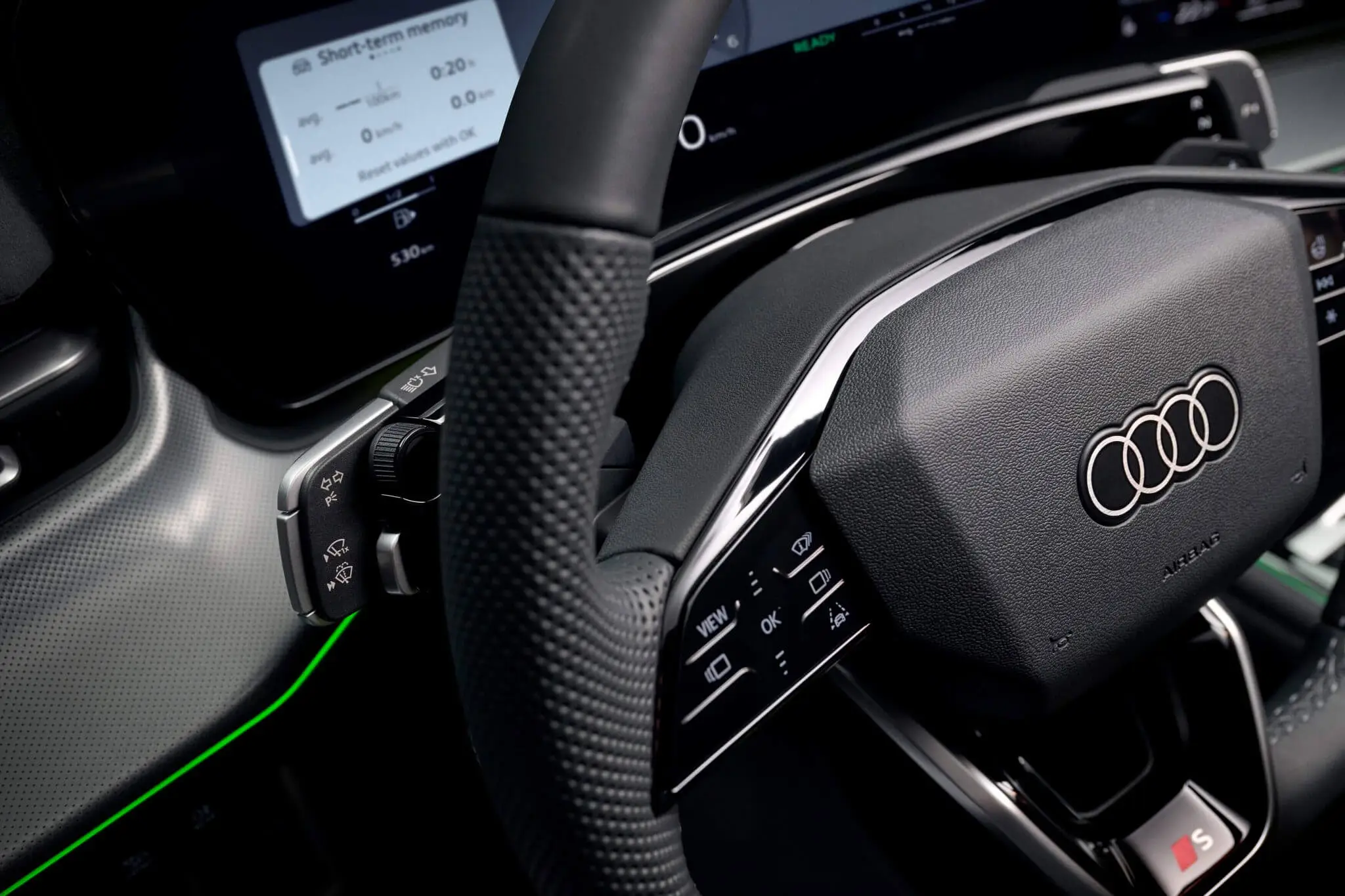 Detalhe do volante do Audi Q3 2026 — logótipo Audi, comandos multifunções, painel de instrumentos digital e iluminação ambiente verde.